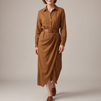 A woman modeling a long-sleeve brown button-front shirt dress with a matching brown belt, standing against a plain warm beige studio background.