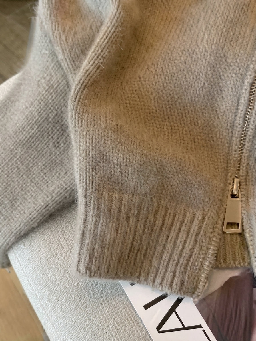 Taupe cropped knit cardigan with a vintage patchwork design, front zipper closure, and dark brown faux-leather collar displayed on a cream armchair.