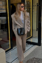 Adele Beige short jacket worn with matching trousers and a white turtleneck, standing in a doorway.