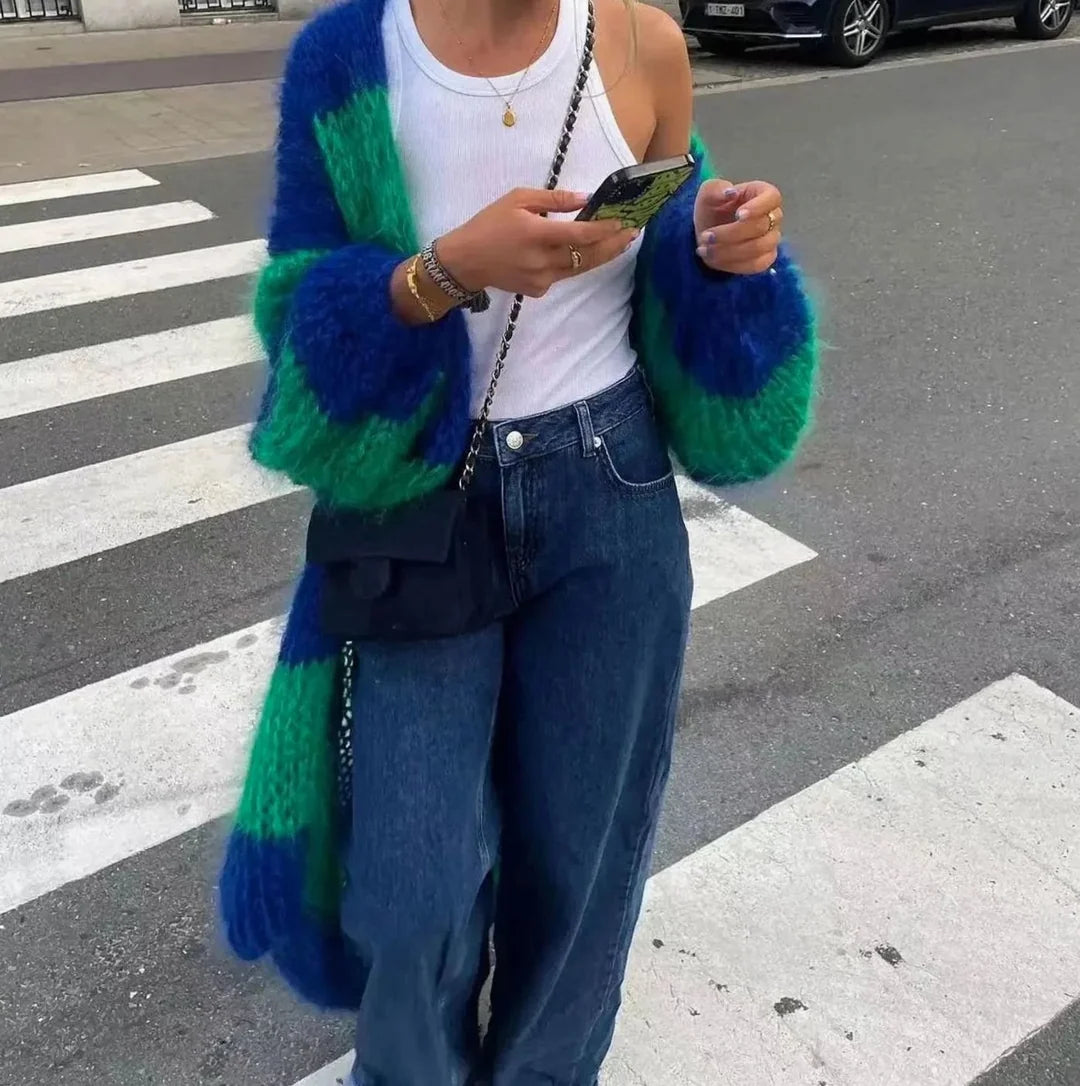 A person standing on a street crosswalk wearing a chunky fuzzy knit cardigan with bold blue and green color-blocked stripes, a white tank top, blue jeans, and a black crossbody bag.