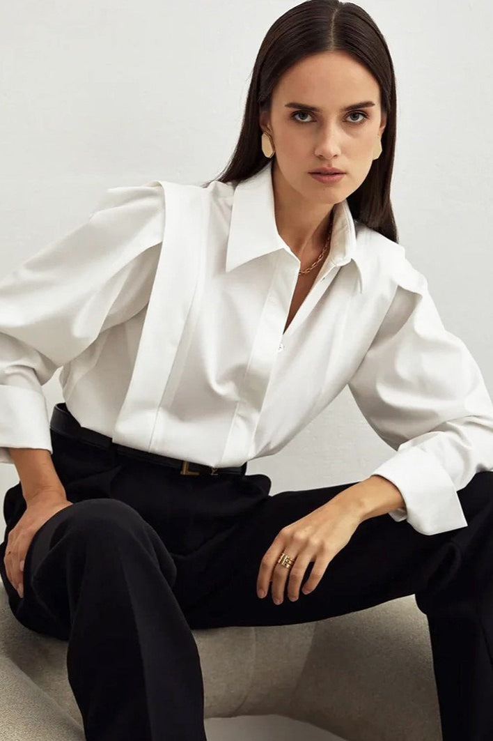 Ariana White button-down shirt styled with a black belt and black trousers while seated, showing the point collar and wide cuffs.