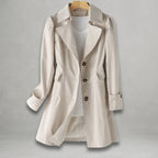 Front view of the Aurora Trench Coat with Belted Notch Collar in beige, featuring wide notched lapels, a double-breasted button front, long sleeves with cuff straps, matching waist belt, and a flared mid-length silhouette. Designed by Bella Hudson.