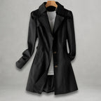 Front view of the Aurora Trench Coat with Belted Notch Collar in black, featuring wide notched lapels, a button-front closure, long sleeves with cuff straps, matching waist belt, and a flared mid-length silhouette. Designed by Bella Hudson.