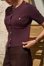 Clara Purple short-sleeve knit cardigan shown from the side with gold buttons and a chest pocket, styled with dark brown trousers.