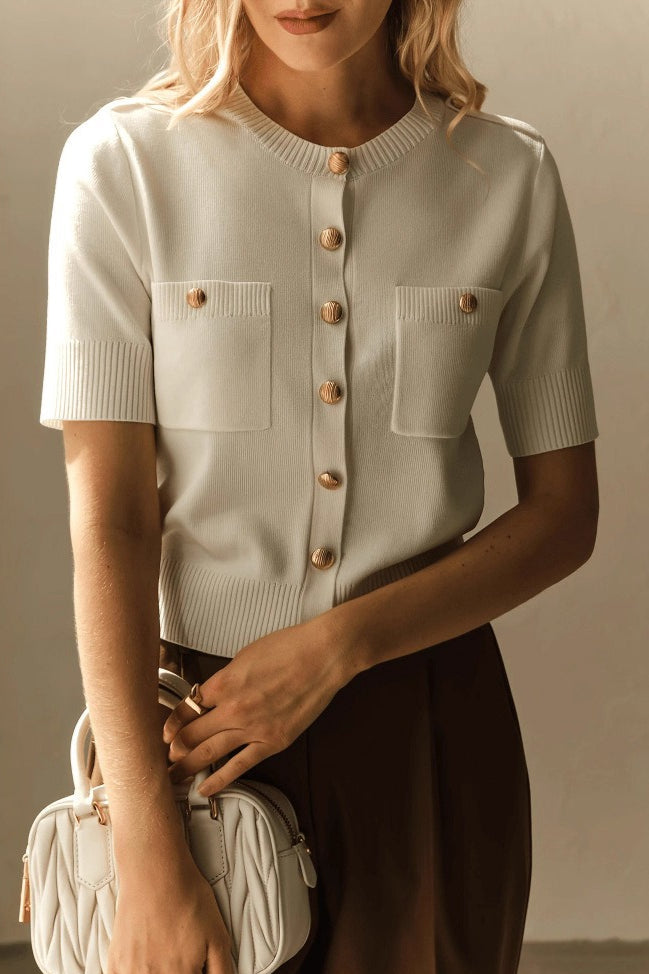 Clara White short-sleeve knit cardigan with gold buttons and patch pockets, styled with brown trousers and a quilted white bag.