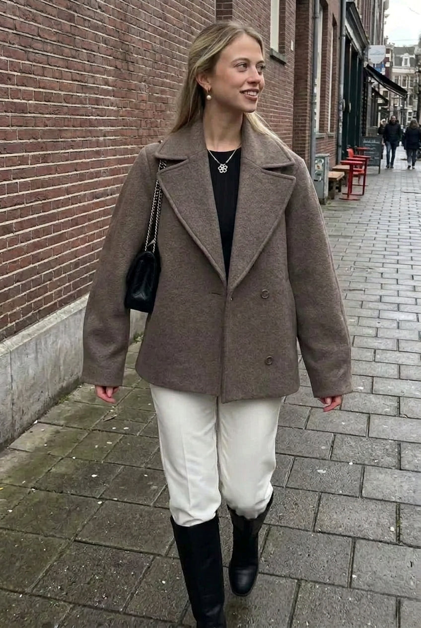 Elise Gray double-breasted lapel coat with wide lapels and front buttons styled with a black top, cream pants and black boots on a city street.