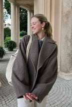 Elise Gray oversized double-breasted lapel coat worn open over a sweater and white shirt, shown from the front under a stone colonnade.