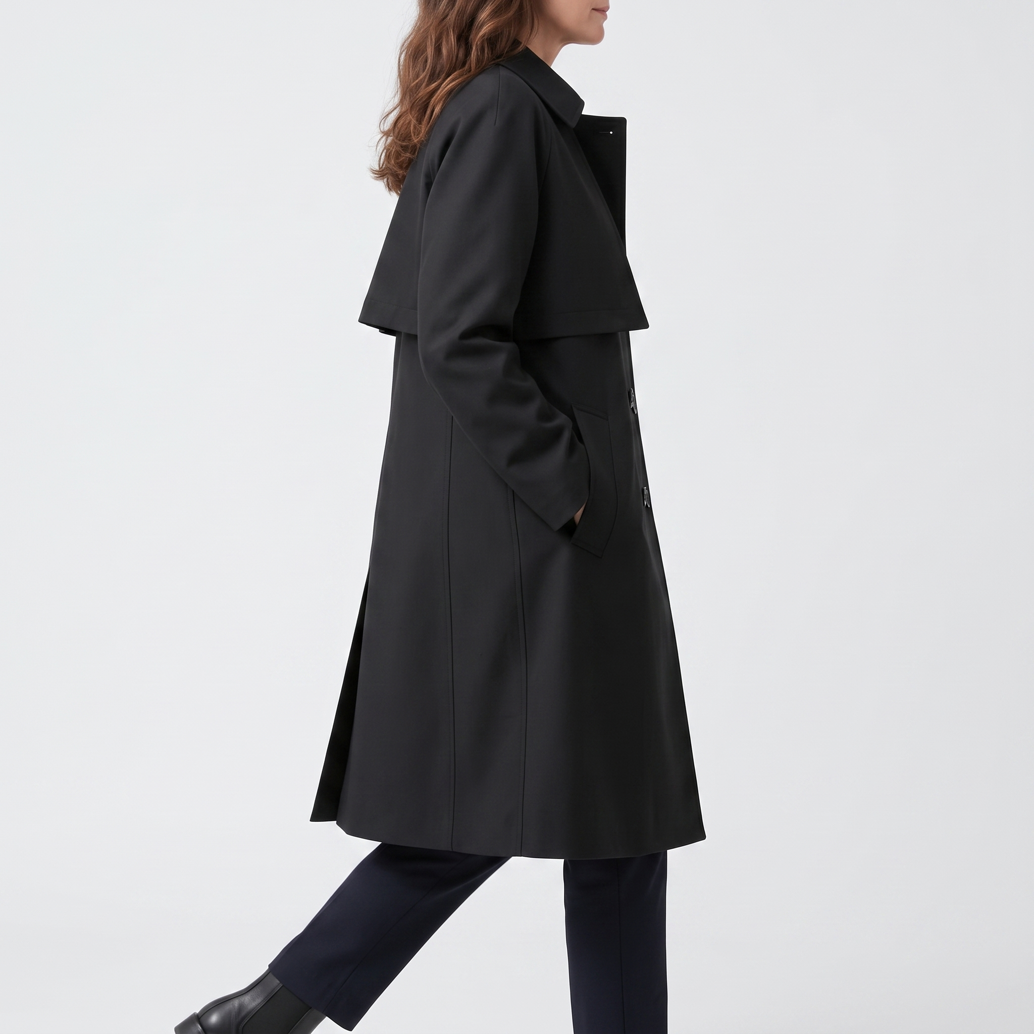 Side view of the Emilia Trench Coat Midi Double-Breasted in black, showcasing the pointed collar, long sleeves, straight cut, side pocket, and midi-length hem. Designed by Bella Hudson.
