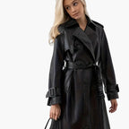 Front view of the Emma Trench Coat Faux-Leather Belted in black, featuring wide notched lapels, a double-breasted front, matching waist belt, long sleeves with buckle cuffs, and a glossy faux-leather finish. Designed by Bella Hudson.