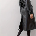 Lower detail view of the Emma Trench Coat Faux-Leather Belted in black, highlighting the belted waist, double-breasted front, smooth faux-leather finish, and straight knee-length hem. Designed by Bella Hudson.
