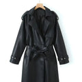 Front product view of the Emma Trench Coat Faux-Leather Belted in black, featuring wide notched lapels, a double-breasted button front, matching tie waist belt, long sleeves with cuff straps, and a knee-length faux-leather silhouette. Designed by Bella Hudson.