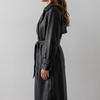 Side view of the Emma Trench Coat Faux-Leather Belted in black, showcasing the wide lapels, belted waist, long sleeves, knee-length hem, and smooth faux-leather finish. Designed by Bella Hudson.