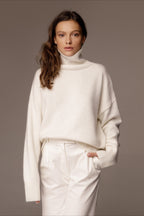 Evelyn White turtleneck sweater with a hand in the pocket of white trousers, showing the ribbed cuffs and dropped shoulders.