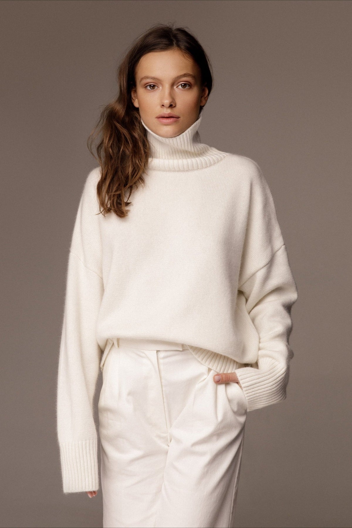 Evelyn White turtleneck sweater with a hand in the pocket of white trousers, showing the ribbed cuffs and dropped shoulders.
