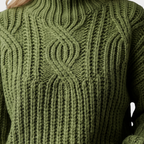 Close-up detail view of the Halvard Sweater Cable-Knit Turtleneck in green, featuring the chunky cable-knit pattern, ribbed turtleneck, and textured knit construction. Designed by Bella Hudson.