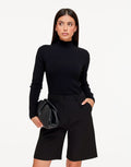 Isolde Black ribbed knit turtleneck sweater styled with black tailored shorts and a black clutch, shown from the front.
