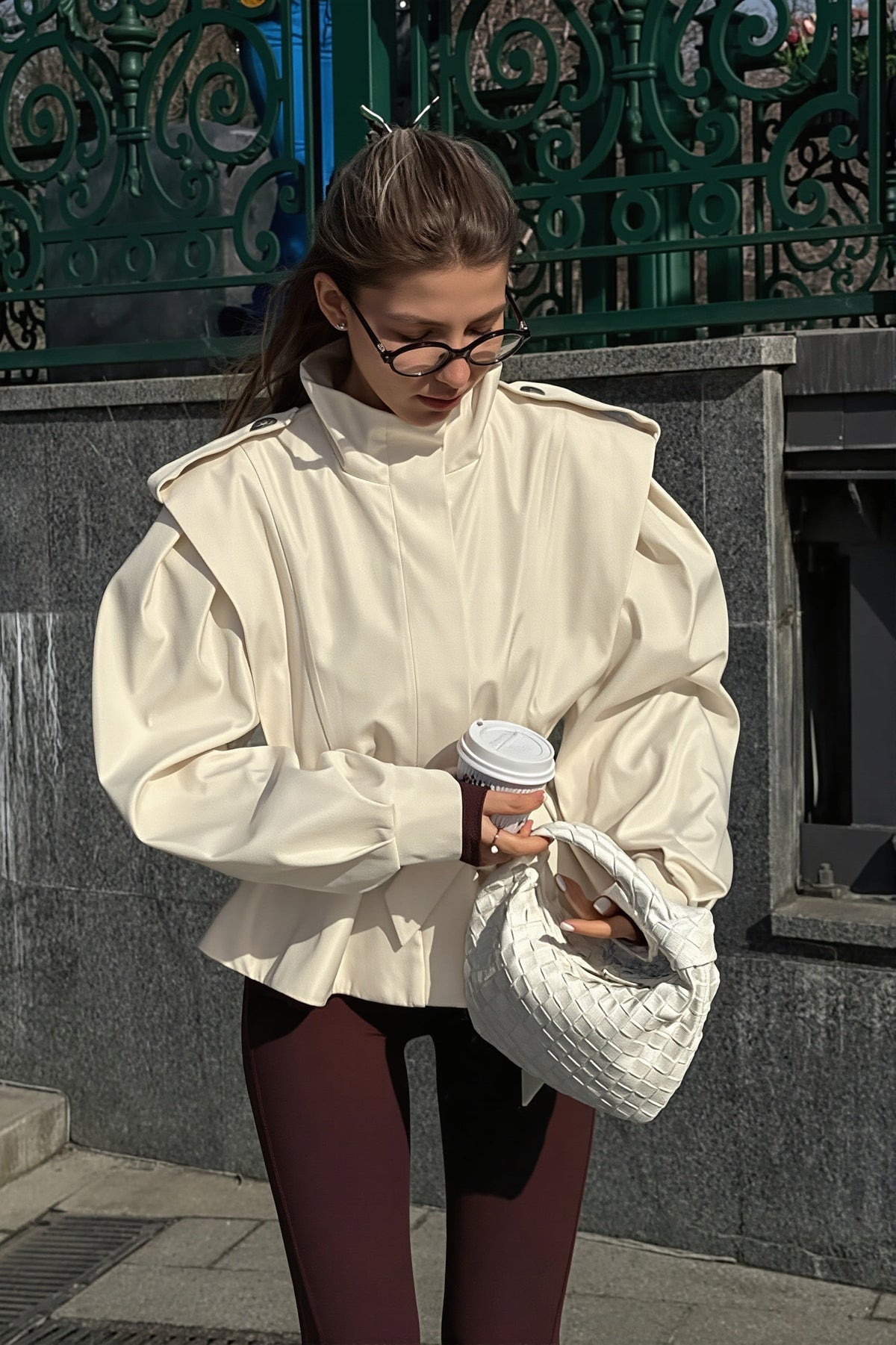 Leona Beige belted peplum jacket shown from the side with structured shoulders and full sleeves while holding a woven bag.