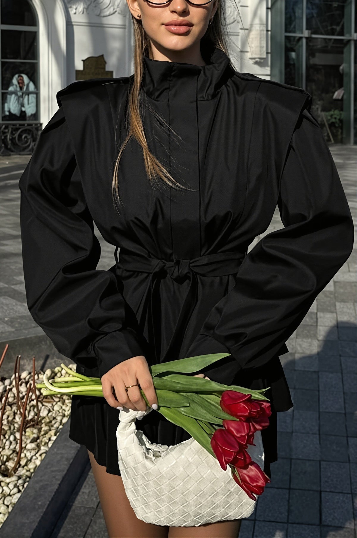 Leona Black belted peplum jacket with stand collar and tied waist, worn with a mini skirt and holding red tulips.