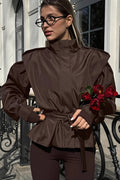 Leona Brown belted peplum jacket with stand collar and structured shoulders, styled with dark leggings and holding red tulips.