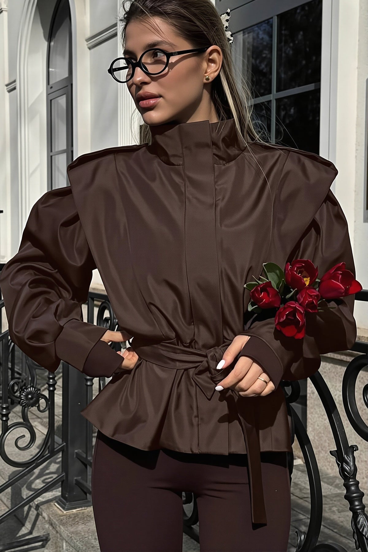 Leona Brown belted peplum jacket with stand collar and structured shoulders, styled with dark leggings and holding red tulips.