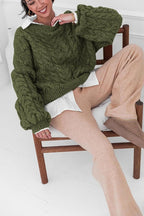 Mila Green cable-knit sweater worn over a white shirt while seated on a wooden chair.