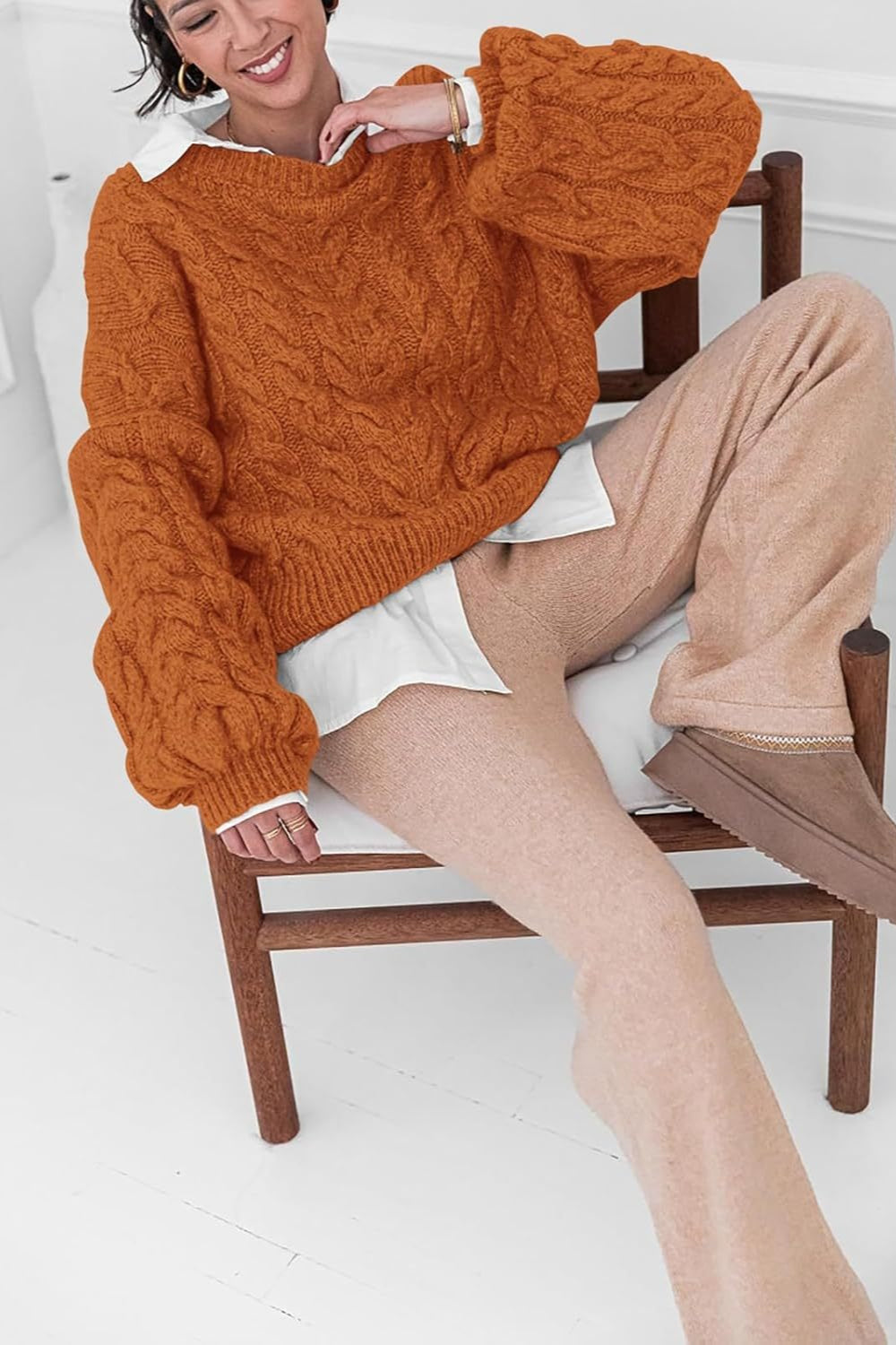 Mila Orange cable-knit sweater worn over a white shirt while seated on a wooden chair.