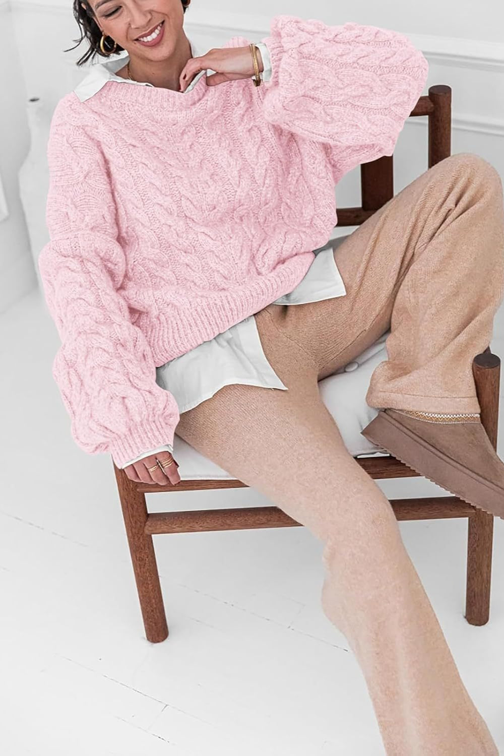 Mila Pink cable-knit sweater worn over a white shirt while seated on a wooden chair.