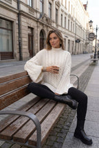 Mila White cable-knit mock-neck sweater styled with black leggings and boots while seated on a bench.