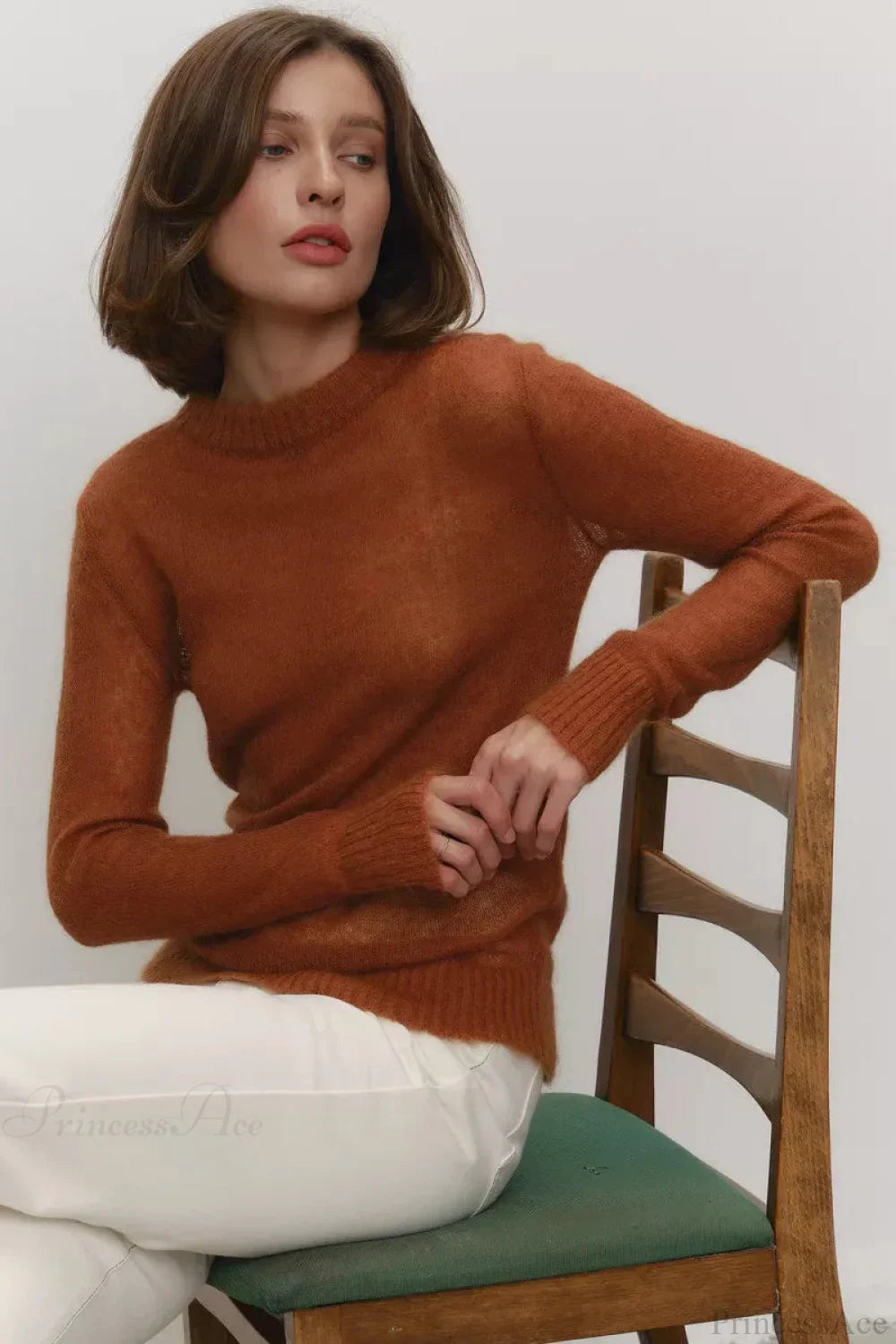 Selena Brown crewneck sweater worn seated on a wooden chair with white pants, showing the long sleeves and ribbed hem.
