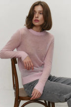 Selena Pink sheer-knit crewneck sweater worn seated on a chair with gray jeans, showing the ribbed cuffs and hem.