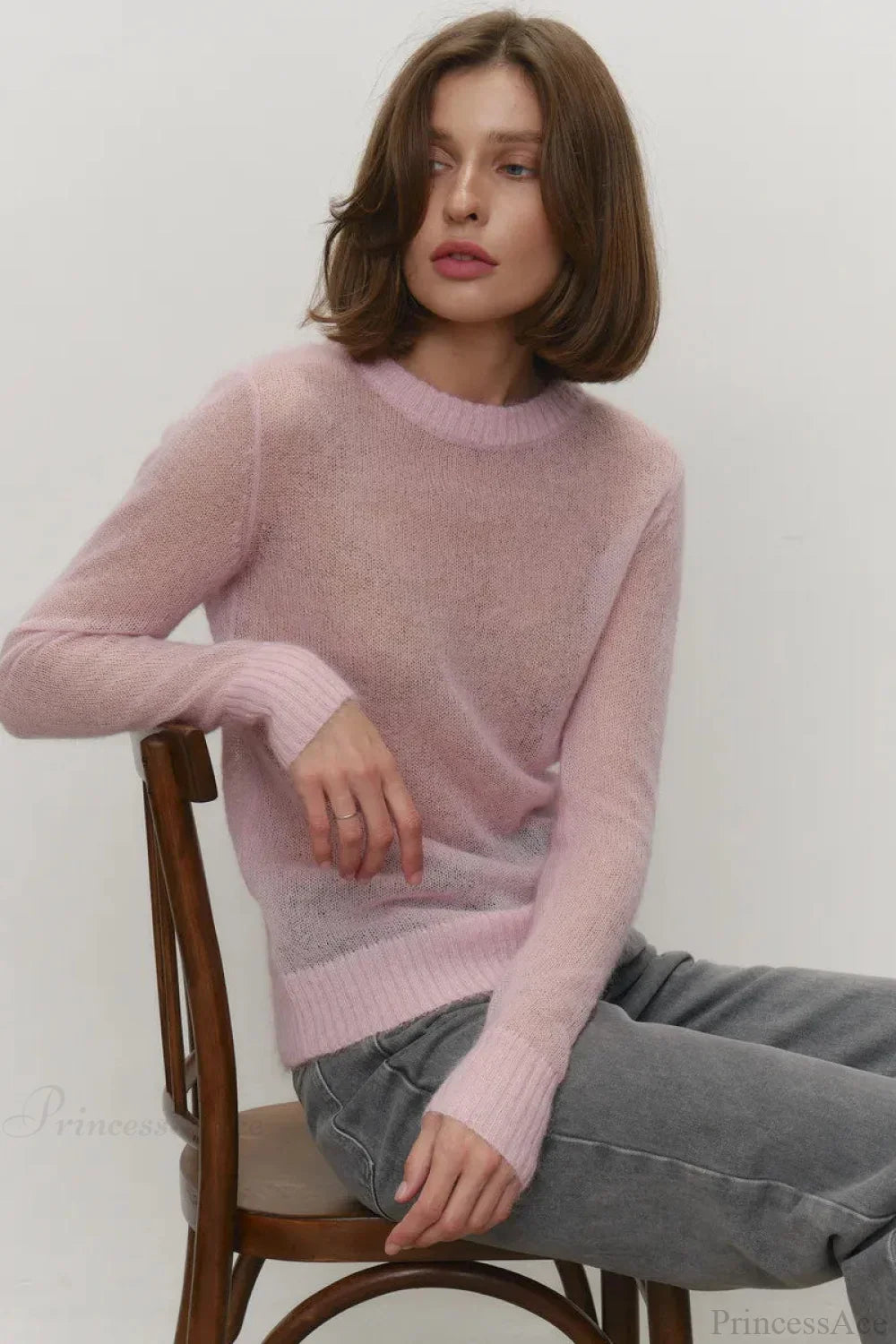 Selena Pink sheer-knit crewneck sweater worn seated on a chair with gray jeans, showing the ribbed cuffs and hem.