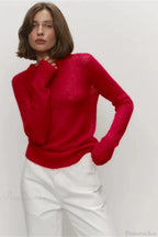 Selena Red sheer-knit crewneck sweater paired with white pants, with one hand raised to the neckline.