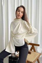 Selene Apricot blouse styled seated on a cane chair, showing the wide flared sleeves and draped front.