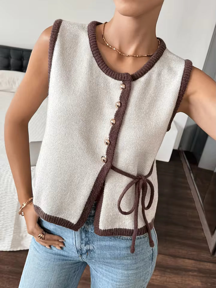 Sophie Beige knit sweater vest with contrast trim, decorative buttons, and side tie, styled with jeans in a seated pose